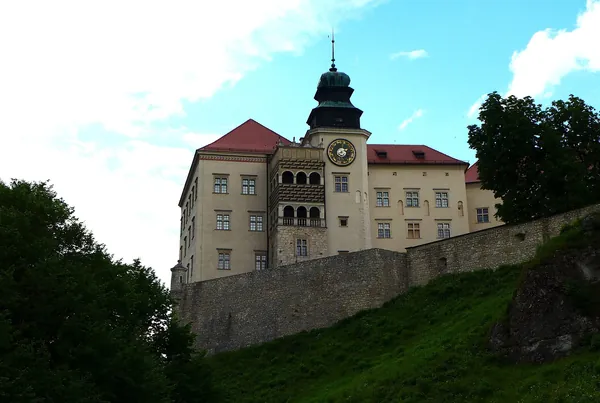 Zamek Pieskowa Skała widziany z dołu — jasny renesansowy zamek z wieżą zegarową i czerwonym dachem na skale, nad zielonym zboczem